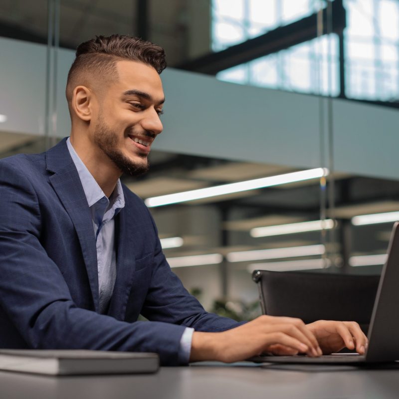 Cheerful young middle eastern businessman sitting at worktable at modern office, typing on notebook keyboard, chatting with business partners, working on marketing research, copy space, side view